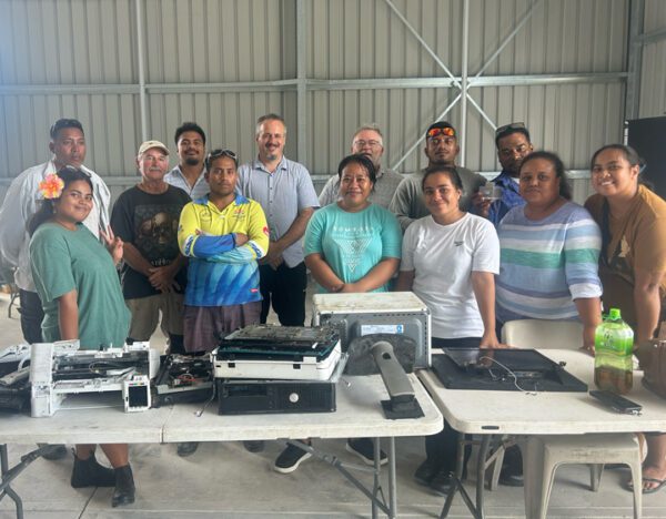 Group photo from Kiribati e-waste training with tables of dismantled printers, computer parts, and electronic components in a workshop.