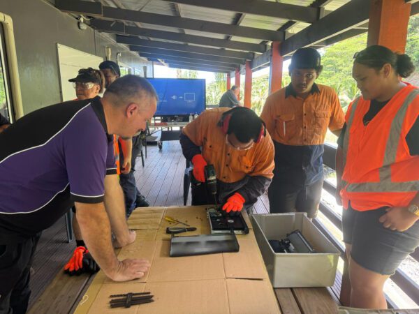 Rarotonga Island e-waste management training session showing participants in safety vests dismantling electronic devices on a table, with tools and components visible.