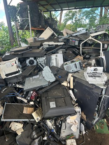 Pile of discarded e-waste under a covered area in Samoa, including old monitors, printers, cables, and appliances for recycling.