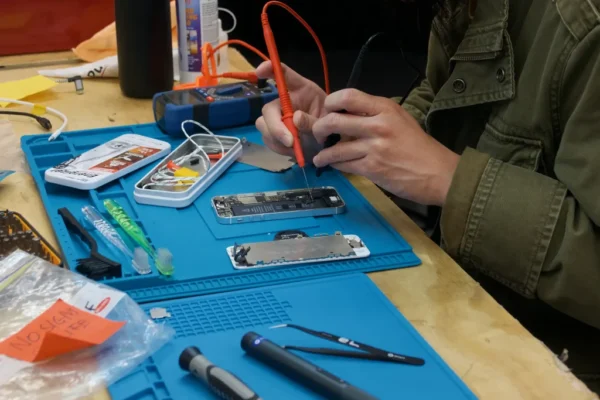 Hands repairing a smartphone on a workbench with tools and wires for The Reconnect Project, promoting e-waste reduction.