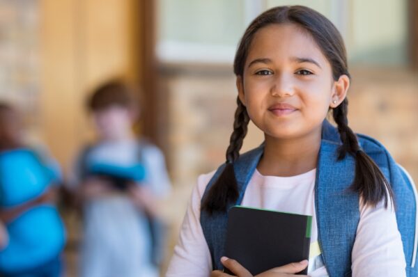 Child with backpack holding a notebook in a school setting, representing The Laptop Initiative for education and digital inclusion.